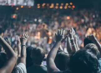 JOHN BARTUS: WHY CONCERTS AND SPORTS ARE SO DARN EXPENSIVE Energetic crowd at a live concert with hands raised and bright lights.
