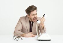MILES TO GO: BY THE POWER VESTED IN ME… Businessman in a beige suit shouting into a vintage telephone, expressing frustration at work.