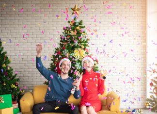 Man and Woman Wearing Santa Hats Sitting on Sofa Popping A Confetti Surrounded By Christmas trees