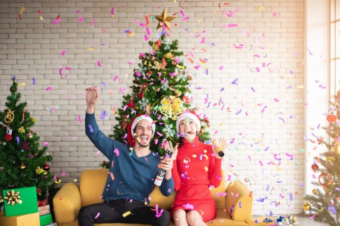 Man and Woman Wearing Santa Hats Sitting on Sofa Popping A Confetti Surrounded By Christmas trees