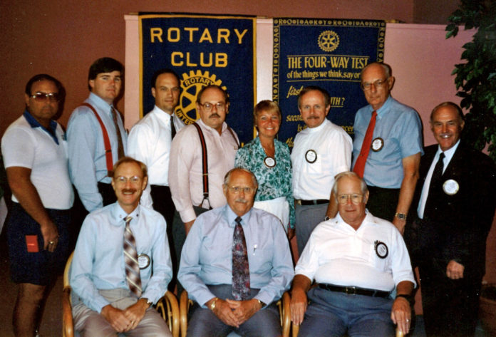 a group of men standing and sitting in front of a rotary club sign