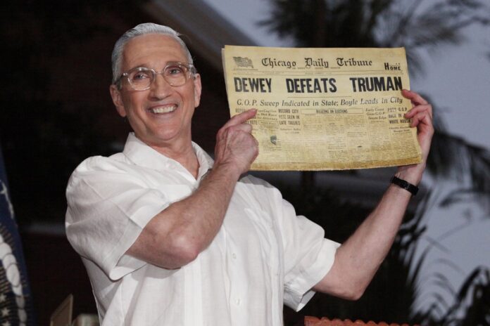 2 Give Em Hell_C Tedesco_1500 Large a man holding up a newspaper in front of an american flag