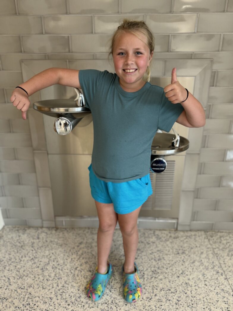 a little girl standing in front of a water fountain giving a thumbs up