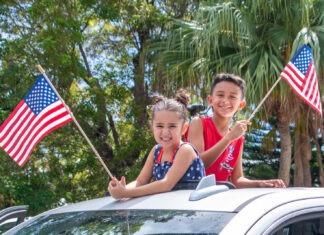 two children waving american flags from the roof of a car