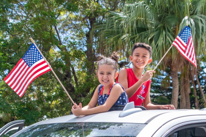 two children waving american flags from the roof of a car