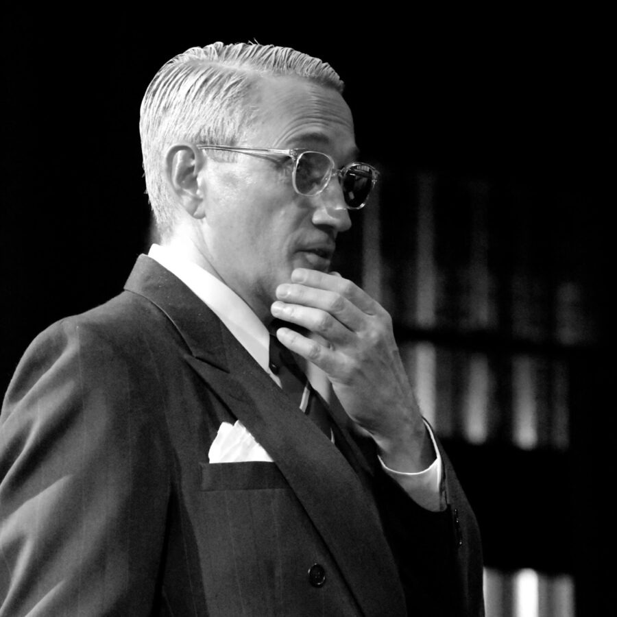 a man in a suit and glasses standing in front of a bookshelf