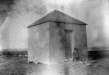 a black and white photo of a man standing in front of a small building