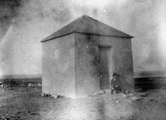 a black and white photo of a man standing in front of a small building