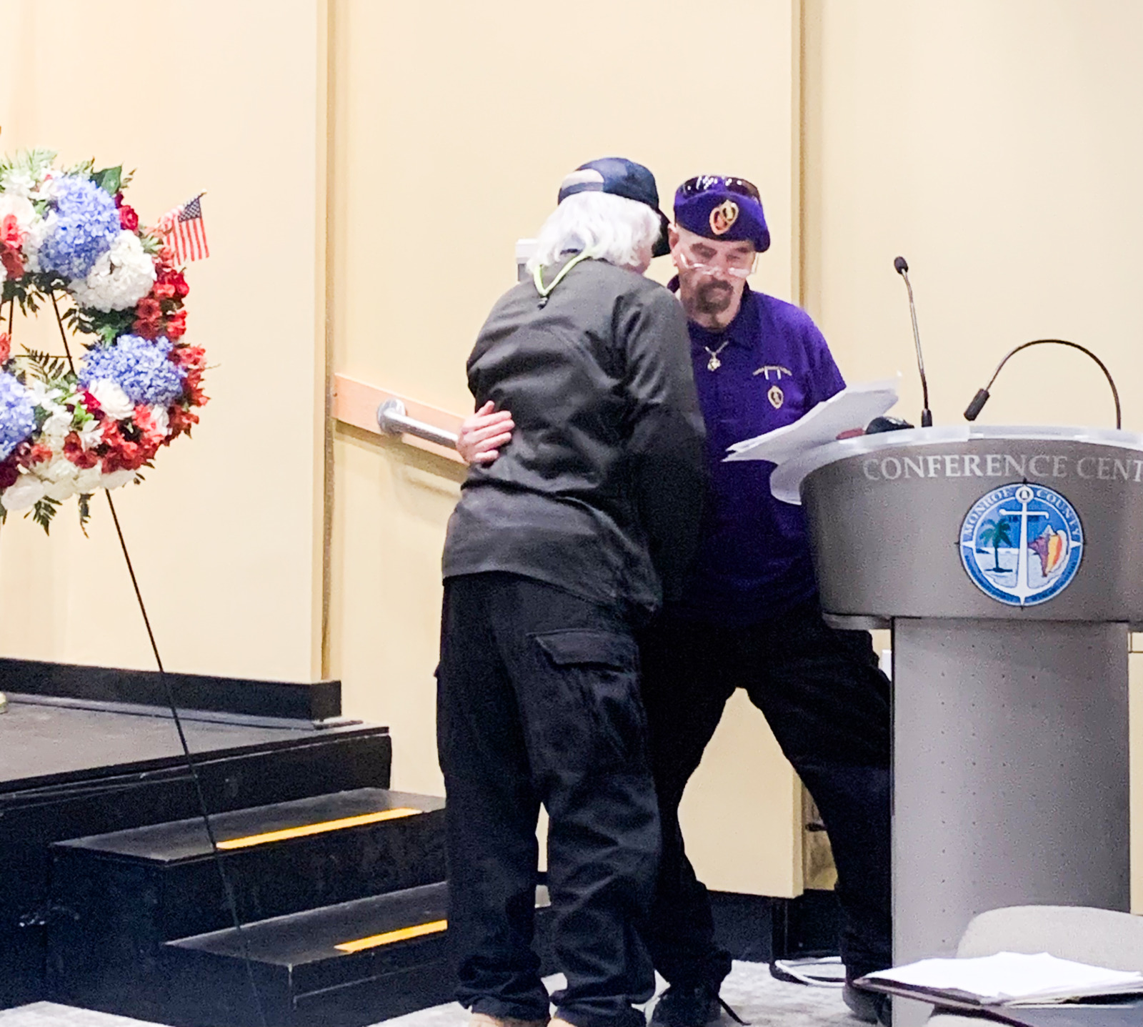 two people standing at a podium with a wreath of flowers behind them