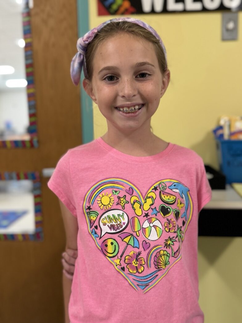 a young girl standing in a classroom wearing a pink shirt