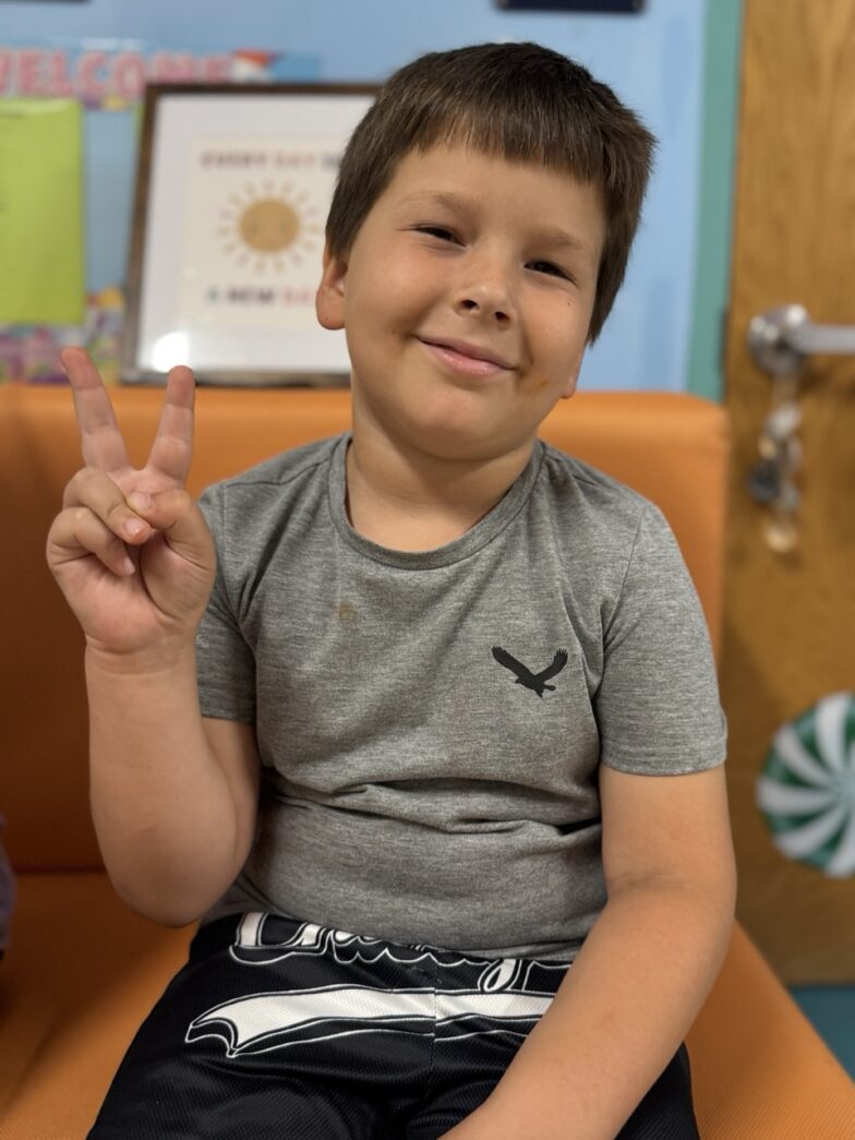 a young boy sitting on a chair making a peace sign