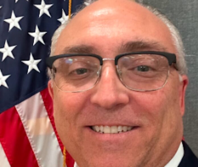 Screenshot a man in a suit and tie in front of an american flag