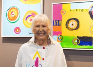 MEET JEAN CARPER, THE LIFE-LOVING WOMAN WHO WROTE ‘DEAD’ a woman standing in front of two paintings