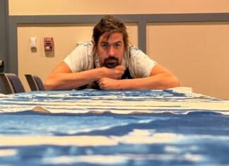 MARATHON CITY HALL’S NEWEST ART GALLERY REFRAMES HISTORY IN ROYAL BLUE a man sitting at a table with his arms crossed